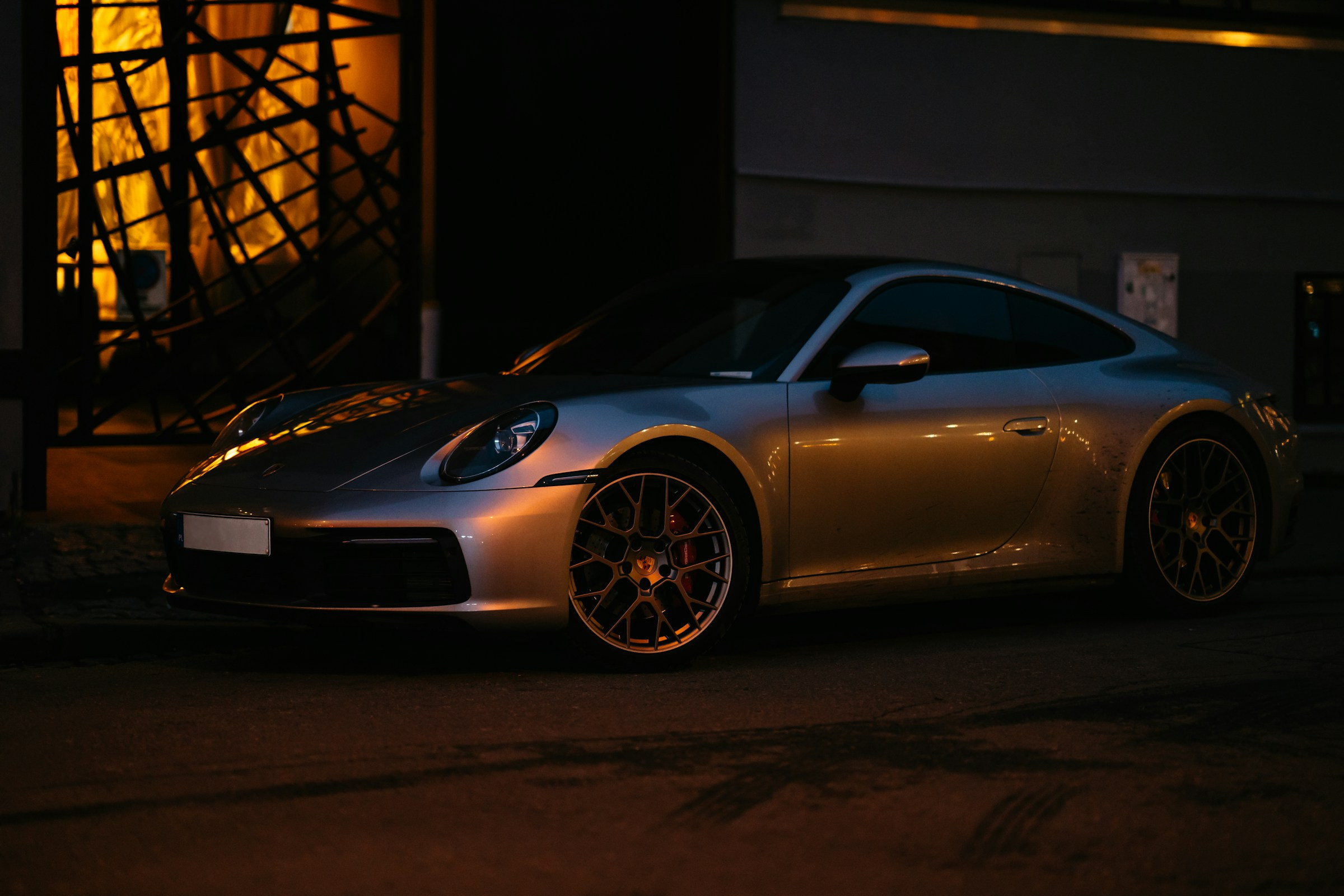 A real silver Porsche photographed at night, with warm light reflected in its paint and detailed alloy wheels; reference photography